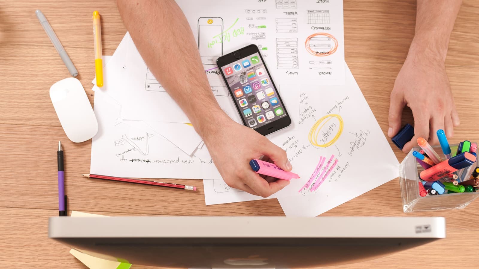 Overhead view of a desk with hand-drawn wireframes, a smartphone and pens, with a hand holding a pink highlighter above the edge of a monitor