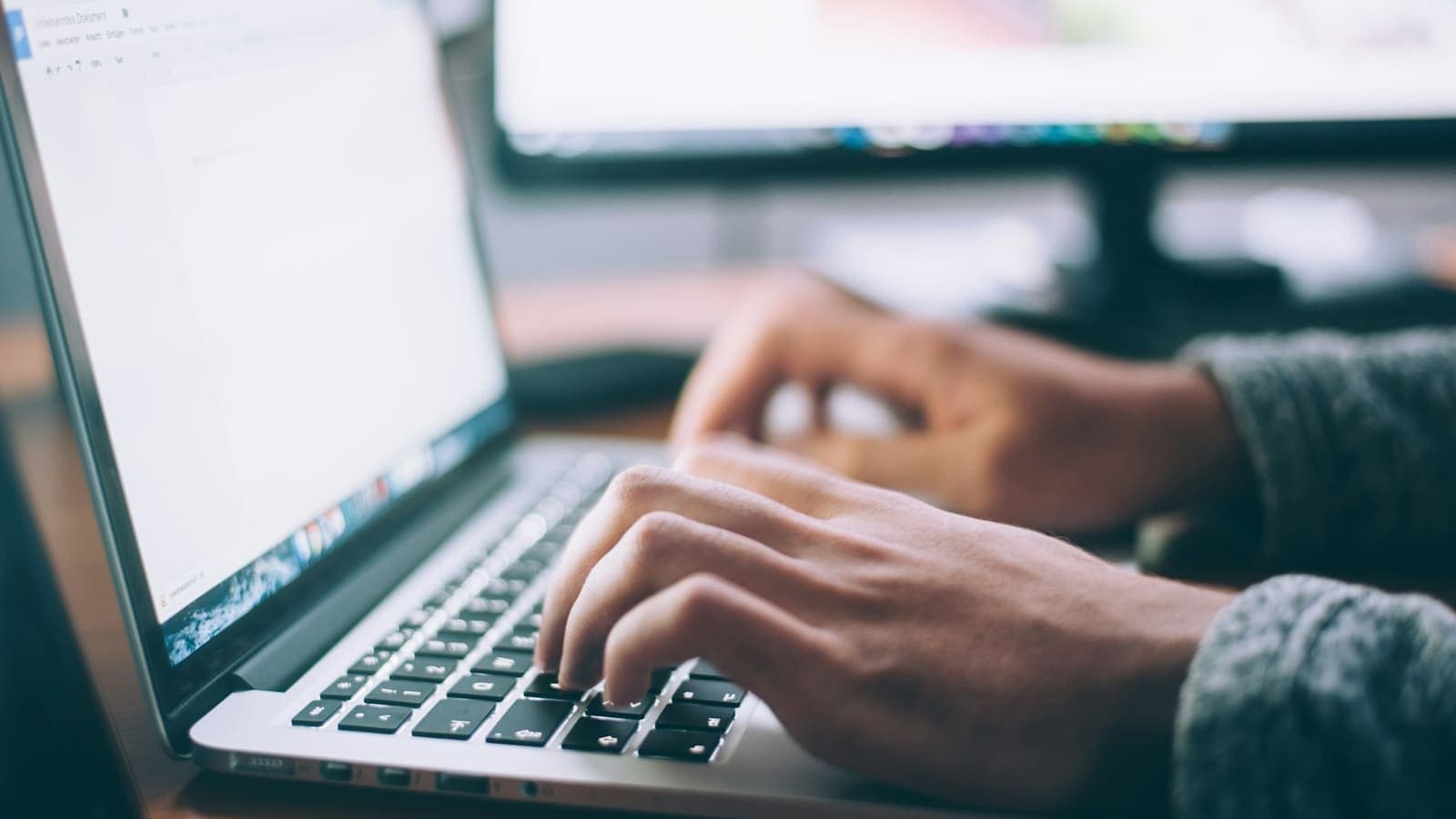 Hands typing on a laptop keyboard with a second monitor blurred in the background